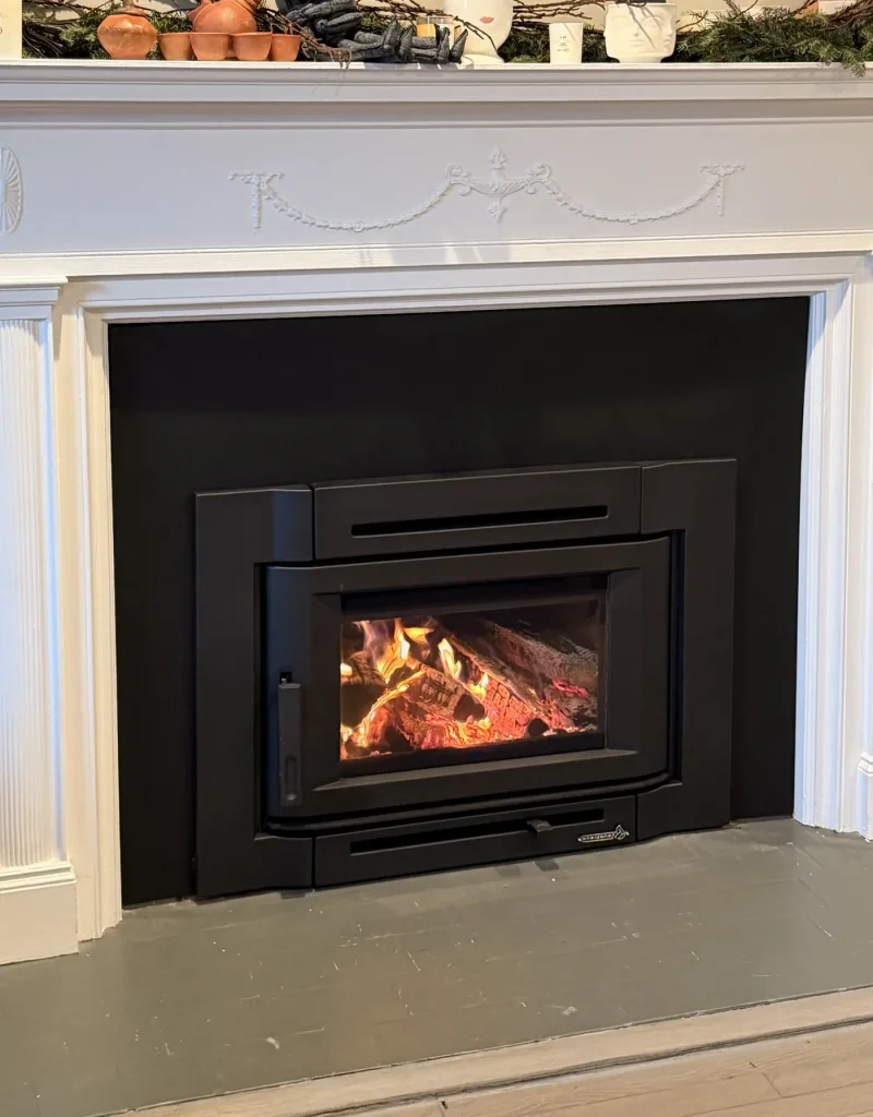 Wood-burning insert with a fire lit, set into a white painted fireplace surround, a Hearthbeats project